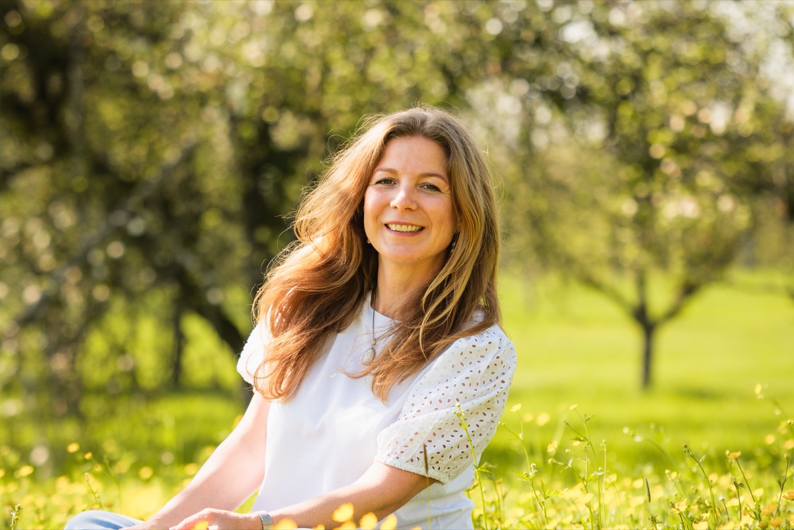 Sarah Veyel sitzt lächelnd in einer Blumenwiese unter Obstbäumen
